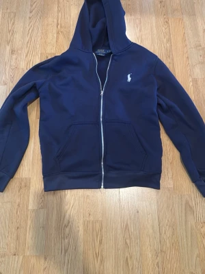 Mörkblå hoodie från Polo Ralph Lauren - Mörkblå hoodie med dragkedja från Polo Ralph Lauren. Klassisk design med huva, två fickor framtill och den ikoniska vita logotypen broderad på bröstet. Tillverkad i mjukt material som känns skönt mot huden. Perfekt för en avslappnad stil. PRISET GÅR ATT DISKUTERA!