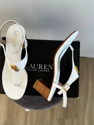 Vita lädersandaler från Lauren Ralph Lauren - Stilrena vita lädersandaler i  krokodilmönster från Lauren Ralph Lauren med guldfärgade RL-detaljer framtill. Sandalerna har en justerbar rem med spänne och en trendig träklack. Tillverkade i ett material med krokopräglad struktur för en lyxig känsla. • Klack på 6 cm och ett justerbart spänne.  • Endast testade men oanvända. I nyskick.  • Passar, beroende på foten, 37.5 -38 (7B).  • Kan mäta & skicka bilder vid intresse.