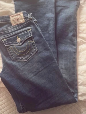 Blå jeans från True Religion - Säljer ett par klassiska blå jeans från True Religion med ikonisk fickdesign och kontrastsömmar. Jeansen har raka ben och detaljerade bakfickor med gul söm och knapp. Perfekta för dig som gillar streetwear och vill ha en tidlös look.