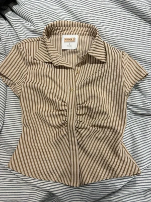 Beige och brunrandig blus från Shein - Kortärmad blus från Shein med beige och bruna vertikala ränder. Modellen har krage, knäppning framtill och rynkade detaljer vid bysten. Tillverkad i ett lätt tyg som känns skönt mot huden. Perfekt för en trendig och avslappnad look.