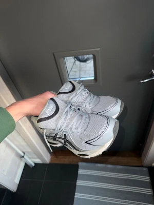 Grå Asics sneakers med svarta detaljer - Säljer ett par grå sneakers från Asics med svarta och silvriga detaljer. Skorna har mesh- och syntetmaterial i ovandelen, klassisk snörning och en vit sula med svarta inslag. Perfekta för dig som gillar sportig och clean stil.