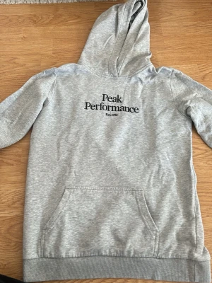 Grå hoodie från Peak Performance - Snygg grå hoodie från Peak Performance med svart logga och text på bröstet. Klassisk känguruficka framtill och ribbade muddar vid ärmslut och nederkant. Perfekt för en avslappnad och sportig stil. Mjuk bomullsblandning och huva.