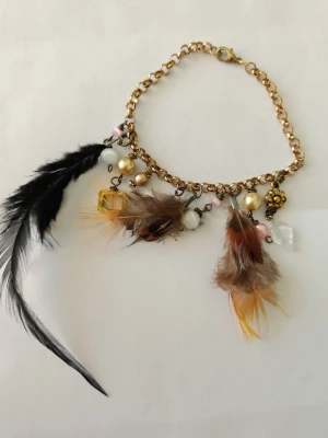 Bohemiskt armband med fjädrar och pärlor - Unikt armband i guldfärgad metall med hängande detaljer som färgglada fjädrar, vita och guldiga pärlor samt små stenar. Armbandet har en chunky kedja och karbinlås. Perfekt för dig som gillar boho-stil och vill sticka ut med accessoarer. Jag kan anpassa längd så det passar