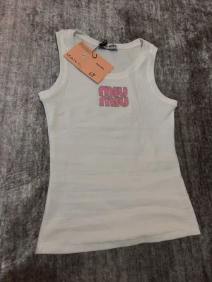 Vit ribbad linnetopp från Miu Miu - Supersnygg vit ribbad linnetopp från Miu Miu med glittrig rosa logga framtill. Toppen är ärmlös och har en tight passform som framhäver figuren. Perfekt för dig som vill ha en trendig och enkel topp med exklusiv känsla.