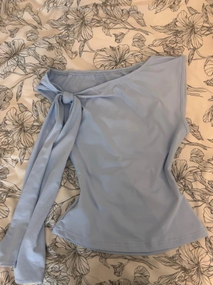 Ljusblå omlottopp med knyt - Snygg ljusblå topp med omlottdesign och knyt vid axeln. Toppen är ärmlös och har en asymmetrisk, oneshoulder-look