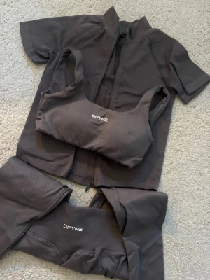 Grått träningsset från DFYNE - Snyggt grått träningsset från DFYNE med sport-bh, kortärmad zip-tröja och tights. Setet är tillverkat i stretchigt, ribbat material som sitter tight och bekvämt. Perfekt för gymmet eller annan träning. Diskret logga på både bh och tights.