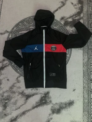 Svart PSG x Jordan vindjacka - Snygg svart vindjacka med huva från Jordan i samarbete med PSG. Jackan har dragkedja framtill, två sidofickor med dragkedja och färgblock i blått och rött över bröstet. PSG-logga och Jumpman-logga på bröstet samt patch nertill. Perfekt för sportig stil.