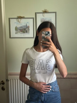 Vit GAP t-shirt med tryck - Säljer en vit t-shirt från GAP med stort tryck 'GAP New York' på bröstet. Klassisk rund halsringning och korta ärmar. Perfekt till jeans eller shorts för en avslappnad stil.