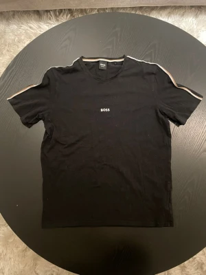 Boss t-shirt - Svart t-shirt från BOSS med korta ärmar och rund hals. Diskret BOSS-logga på bröstet och snygga ränder längs axlarna. Perfekt för en stilren och avslappnad look.