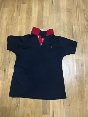 Mörkblå pikétröja från Ralph lauren - Snygg mörkblå pikétröja från Ralph Lauren med röd krage och knappslå. Ikonisk logga broderad på bröstet och tydlig märkning i nacken. Perfekt för en stilren och sportig look. Storlek M men passar även S.