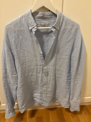 Ljusblå linneskjorta regular fit - Säljer en ljusblå skjorta i 100% linne med regular fit. Skjortan har button down-krage, långa ärmar och vita knappar framtill. Perfekt för en avslappnad men ändå stilren look.