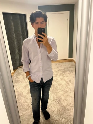 Ljuslila skjorta från Ralph Lauren - Säljer en stilren ljuslila skjorta från Ralph Lauren med klassisk button-down krage och broderad logga på bröstet. Skjortan har långa ärmar och knappar framtill. Perfekt till jeans eller chinos för en clean look.