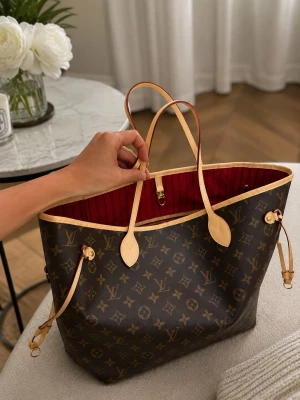 Louis Vuitton Neverfull Monogram Tote - Snygg och rymlig handväska från Louis Vuitton i klassiskt monogrammönster. Väskan har bruna och beige färger med röda detaljer på insidan, långa handtag och sidoremmar. Tillverkad i canvas med läderdetaljer. Perfekt för dig som gillar lyxig och tidlös design.