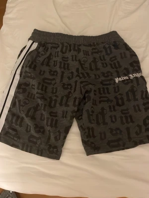 Palm Angels camo shorts med tryck - Säljer ett par gråa shorts från Palm Angels med svart camo- och bokstavstryck. De har elastisk midja och en vit sidoremsa för extra stilpoäng. Perfekta för dig som vill sticka ut med streetwear-vibe.