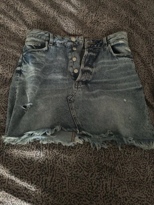 Kort jeanskjol med fransig kant från H&M  - Säljer en kort jeanskjol i blå denim med slitningar och fransig nederkant. Kjolen har knappar framtill och klassiska fickor både fram och bak. Perfekt för dig som gillar en avslappnad och trendig stil.