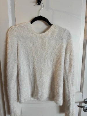 Vit fluffig stickad tröja med knyt - Mysig vit stickad tröja med fluffig textur och lång ärm. Tröjan har rund halsringning fram och en snygg detalj med knyt i nacken.