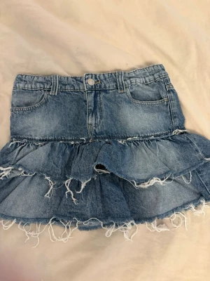 jeanskjol med volange - Säljer en blå jeanskjol med volangdetaljer och råa, fransiga kanter. 
