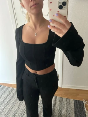 Svart croppad långärmad topp - Snygg svart croppad topp med långa ärmar och fyrkantig urringning. Toppen har en korsettliknande passform som framhäver midjan och ger en trendig look. Perfekt att styla med höga byxor eller kjol för en cool outfit.
