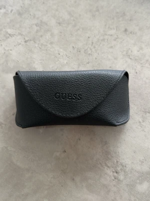Svart solglasögonfodral Guess - Snyggt svart solglasögonfodral från Guess i ett stilrent och klassiskt utförande. Fodralet är gjort i ett strukturerat material som liknar skinn och har en enkel knappstängning. Perfekt för att skydda dina solglasögon i väskan.