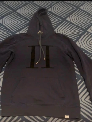  hoodie Les Deux - Snygg mörkblå hoodie från Les Deux med stor svart II-logga på bröstet. Klassisk huva med dragsko och känguruficka framtill. Tillverkad i mjukt material som känns skönt mot huden. Perfekt för en avslappnad och trendig look.