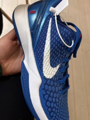 Helt nya kobe 6or  - Säljer helt nya oanvända blå Nike Kobe 6or basketsskor med vit sula och stort vitt Swoosh på sidan. Ovandelen har en texturerad, reptil-liknande yta och blå skosnören. Skon har röda detaljer och Kobe-loggan på plösen. Perfekt för dig som vill sticka ut på planen.