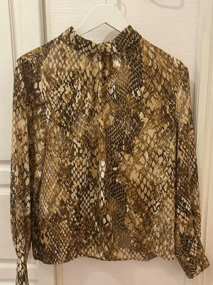 Blus H&M - Snygg blus med ormmönster i bruna och beige toner. Tunt och lätt material som ger en luftig känsla. Perfekt för dig som vill sticka ut med ett trendigt mönster. Aldrig använd, storlek 40 💫