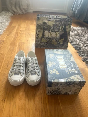 Dior gråvita låga sneakers - Snygga låga sneakers från Dior i gråvitt med klassiskt mönster och tydlig logga på tungan. Skorna har vita snören, gummisula och är tillverkade i textil och läderdetaljer. Perfekta för dig som vill ha en exklusiv touch på din outfit.