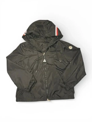 Svart vindjacka från Moncler - Snygg svart vindjacka från Moncler med huva och dragkedja framtill. Jackan har Moncler-logga på ärmen och vita samt röda detaljer på huvan. Tillverkad i glansigt, lätt material och har elastiska muddar vid ärmslut och nederkant.