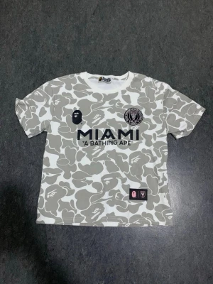 Vit camo t-shirt MIAMI x BAPE - Säljer en limited edition MIAMI x A BATHING APE t-shirt med grått camomönster och stora tryck både fram och bak. T-shirten har korta ärmar och är tillverkad i ett mjukt material, perfekt för dig som gillar streetwear och unika samarbeten.