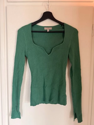 Grön ribbstickad tröja med v-ringning - Snygg grön ribbstickad tröja med lång ärm och djup v-ringad hals. Tröjan har en figurnära passform och är tillverkad i ett mjukt, stretchigt material. Perfekt att styla med jeans eller kjol för en trendig look.