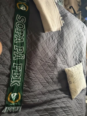 Grön FBK supporterscarf - Mörkgrön halsduk med vit text 'SOPA PÅ FBK' och FBK Karlstad-logga i båda ändar. Klassisk supporterscarf med fransar i ändarna, perfekt för att visa ditt stöd på matchen eller i vardagen.