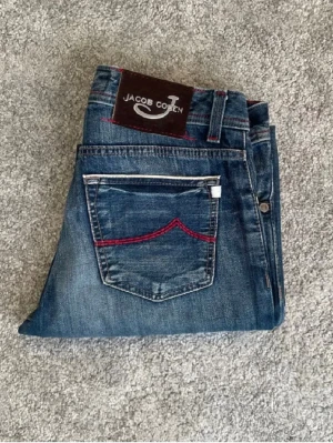 Jacob cohen jeans limited edition 688 - Snygga blå jeans från Jacob Cohen med röda sömmar och detaljer på fickorna. Klassisk femficksmodell med knappar i gylfen och patch med logga bak. Jeansen har en rak passform och är tillverkade i denim. 100% bomull textil lim i anklen på byxorna syns... 