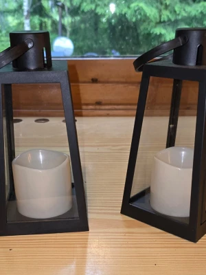 Svarta lyktor med LED-ljus - Två stilrena svarta lyktor i metall och glas med vita LED-ljus inuti. Perfekta som dekoration både inne och ute. Lyktorna har handtag upptill för enkel flytt och ger mysig belysning utan öppen låga. 20 cm höga