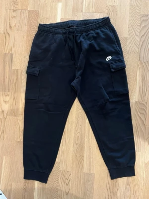 Svarta cargo mjukisbyxor från Nike - Säljer ett par svarta mjukisbyxor från Nike med cargo-fickor på sidorna och dragsko i midjan. Byxorna har ribbade muddar vid bensluten och är tillverkade i mjukt sweatshirtmaterial. Perfekta för en avslappnad och sportig stil.