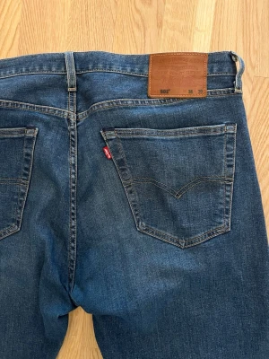 Levi's 501 blå jeans - Klassiska blå Levi's 501 jeans i rak modell med fem fickor och bruna sömmar. Jeansen har en ikonisk läderpatch bak i midjan och röd Levi's-tagg på bakfickan. Tillverkade i slitstark denim, perfekta för en avslappnad och tidlös stil.