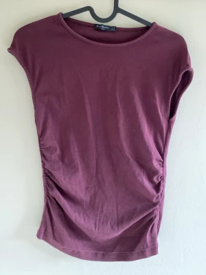 Vinröd topp från Stradivarius - Säljer en vinröd topp från Stradivarius i mjuk bomull. Toppen har rund halsringning, ärmlös design och snygg rynkning på ena sidan som ger en cool twist. Perfekt att styla med jeans eller kjol för en trendig look.