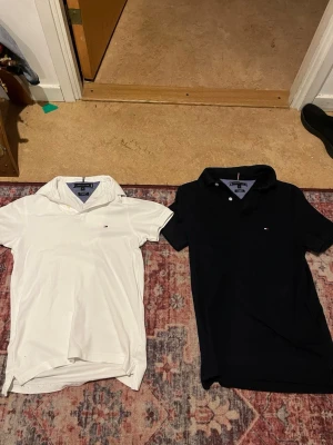 Två pikétröjor från Tommy Hilfiger - Säljer två klassiska pikétröjor från Tommy Hilfiger i storlek XS. En är vit och en är svart, båda med liten logga på bröstet och krage. Slim fit-modell i premium pique-bomull som ger en snygg och bekväm passform.