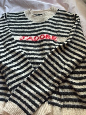 Randig stickad tröja med text - Säljer en svartvit randig stickad tröja med texten 'J'ADORE' i rött framtill. Tröjan har rund halsringning och ribbade muddar vid ärmslut och nederkant. Mysigt material och avslappnad passform, perfekt för en trendig vardagslook.