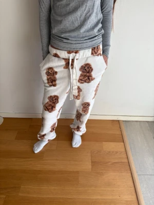 Vita mjukisbyxor med hundmotiv - Supermysiga vita loungewear-byxor med brunt hundmotiv över hela tyget. Byxorna har resår i midjan, dragsko och stora fickor på sidorna. Perfekta för chill dagar hemma och riktigt sköna tack vare det fluffiga materialet.