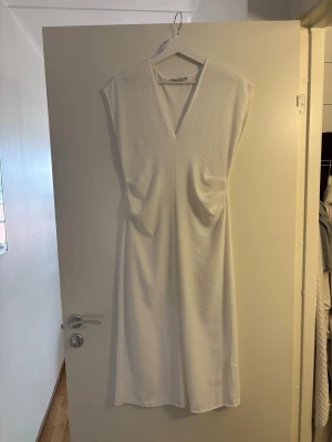 Vit klänning med v-ringning - En stilren vit klänning med djup v-ringning och ärmlös design. Klänningen har en rak passform och är tillverkad i ett mjukt tyg, perfekt för varma dagar. Enkel och minimalistisk look som passar till många tillfällen.