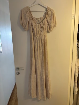 Ljusbeige långklänning med puffärm - Superfin ljusbeige långklänning med puffärmar och smockad struktur. Klänningen har omlott-detalj i ryggen och en luftig, flowy passform. Perfekt för sommarens alla tillfällen och ger en romantisk vibe.