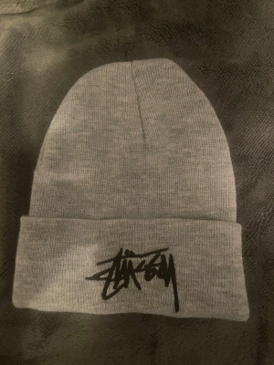 Grå Stüssy mössa med logga - Säljer en stilren grå mössa från Stüssy med den klassiska svarta loggan broderad framtill. Mössan är ribbstickad och har uppvikt kant för extra streetwear-vibe. Perfekt accessoar för att lyfta din outfit under kyliga dagar.