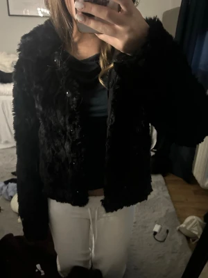 Svart kort pälsjacka - Säljer en svart, fluffig och kort pälsjacka med krage. Jackan har en mjuk fuskpälsstruktur och stängs med knappar framtill. Perfekt för att hålla värmen under kalla dagar och ger en trendig look till din outfit.