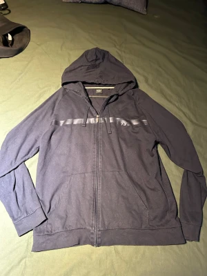 Mörkblå hoodie från BOSS - Snygg mörkblå hoodie från BOSS med dragkedja framtill och huva med dragsko. Har en diskret BOSS-logga på bröstet och en stilren rand över bröstet. Tillverkad i mjukt bomullsmaterial och har två fickor framtill. Perfekt för en avslappnad look.