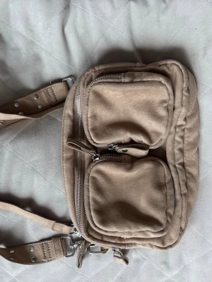 Beige axelremsväska Gina tricot - Snygg beige handväska i mjukt mockaliknande material med två framfickor med dragkedja och justerbar axelrem med nitar. Perfekt för dig som gillar praktiska detaljer och stilren design.