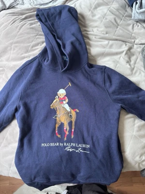 Marinblå hoodie Polo Bear Ralph Lauren - Snygg marinblå hoodie från Polo Ralph Lauren med stor Polo Bear-tryck framtill där björnen rider häst och spelar polo. Klassisk huva och mjukt material som känns skönt mot huden. Perfekt för dig som gillar streetwear och ikoniska prints.