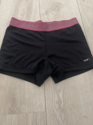 Svarta träningsshorts från SOC - Säljer ett par svarta träningsshorts från SOC med bred elastisk midjeresår i grått och rosa. Shortsen har en tight passform och är tillverkade i ett stretchigt syntetmaterial som passar perfekt för aktiva dagar.