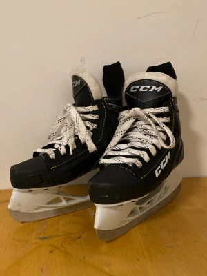 Svarta CCM skridskor med vita detaljer - Säljer ett par svarta CCM skridskor med vita snören och vita detaljer. Skridskorna har robust konstruktion, vadderad plös och klassisk CCM-logga på sidan och plösen. Sulan är vit och skenan i metall. Perfekta för isaktiviteter!