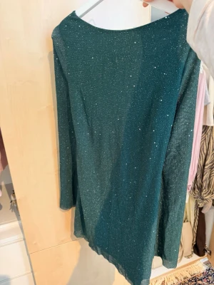 Mörkgrön glittrig kortklänning - Snygg mörkgrön kortklänning med glittrigt tyg och lång ärm. Klänningen har en öppen rygg med knytband upptill och ett lätt draperat fall baktill. Perfekt för dig som vill sticka ut med lite extra glitter. Nypris 399 mitt pris 100