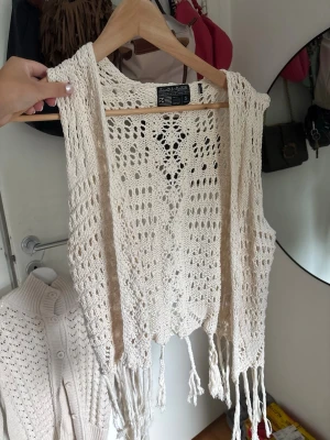 Beige stickad väst med fransar - Säljer en beige stickad väst med virkade detaljer och fransar längs nederkanten. Västen är öppen framtill och har ett bohemiskt uttryck som passar perfekt till lager på lager. Superfin att styla över en enkel topp eller klänning.